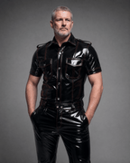 Men’s high shine black PVC fetish shirt with red stitching, short sleeves, button-up front, and chest pockets, worn by a male model in a studio setting.